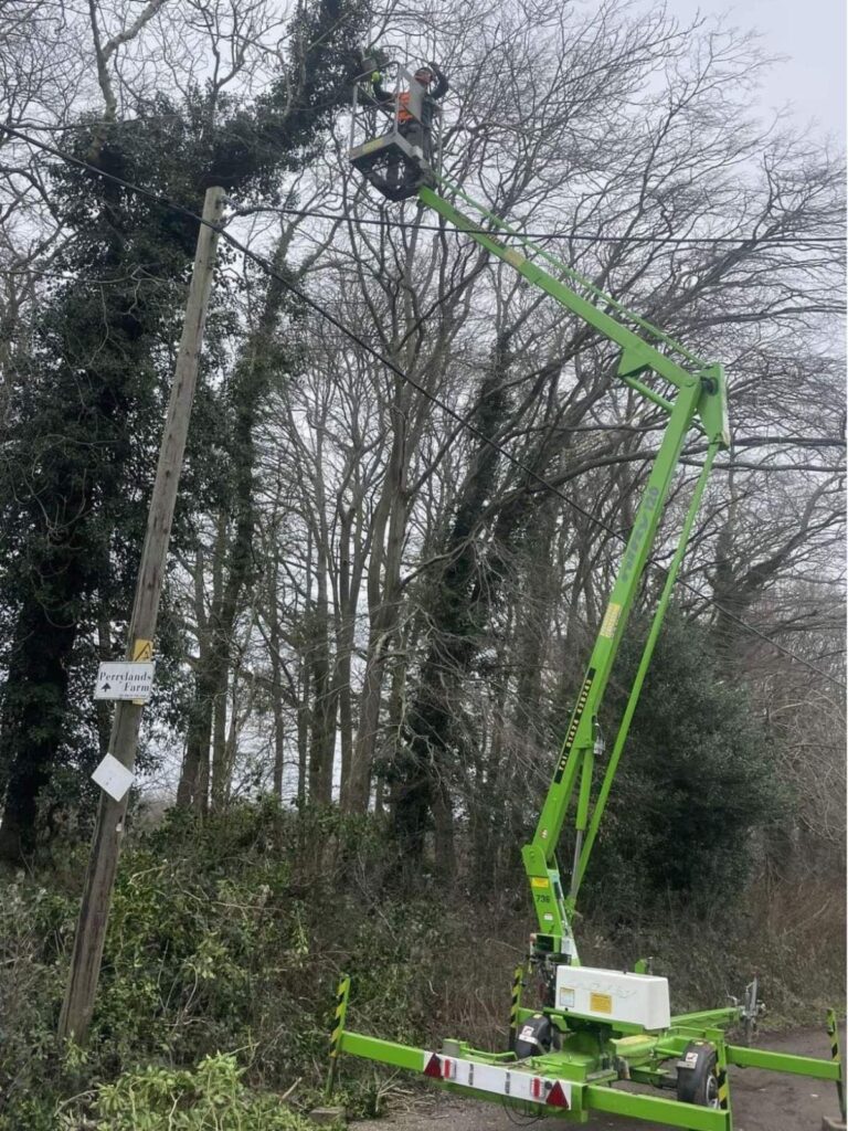  a bright green cherry picker next to a line of trees with a man in the extended cherry picker basket