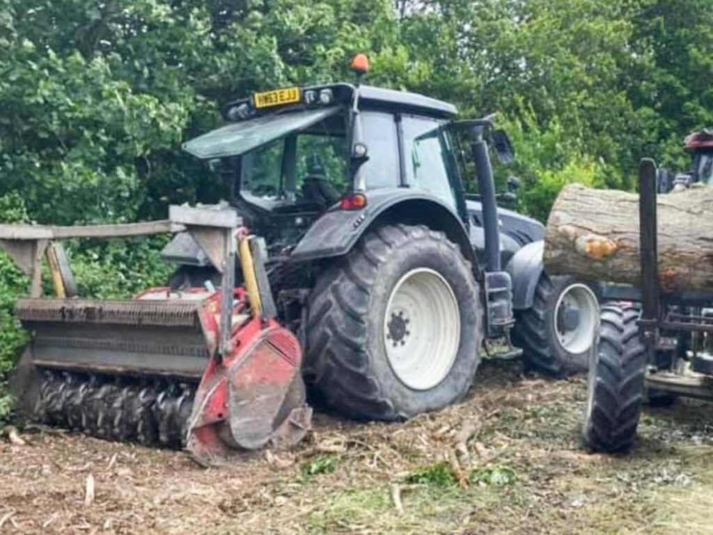 a tractor with a mulcher on the back by a hedge and tree line